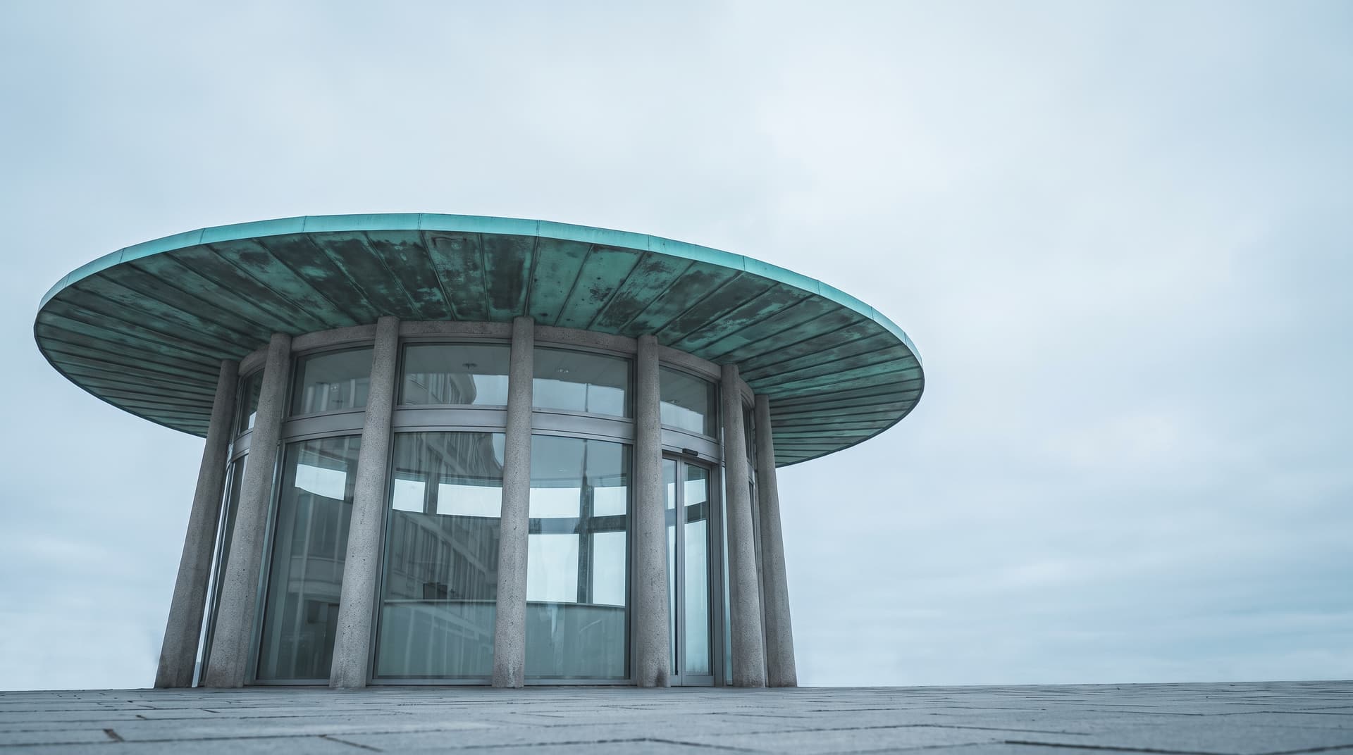 Runder Auskunfts-Pavillon aus Glas und Beton von unten, darüber ein flaches Kreisdach aus grün patiniertem Kupfer – Sinnbild für einen Ort, an dem Fragen beantwortet werden