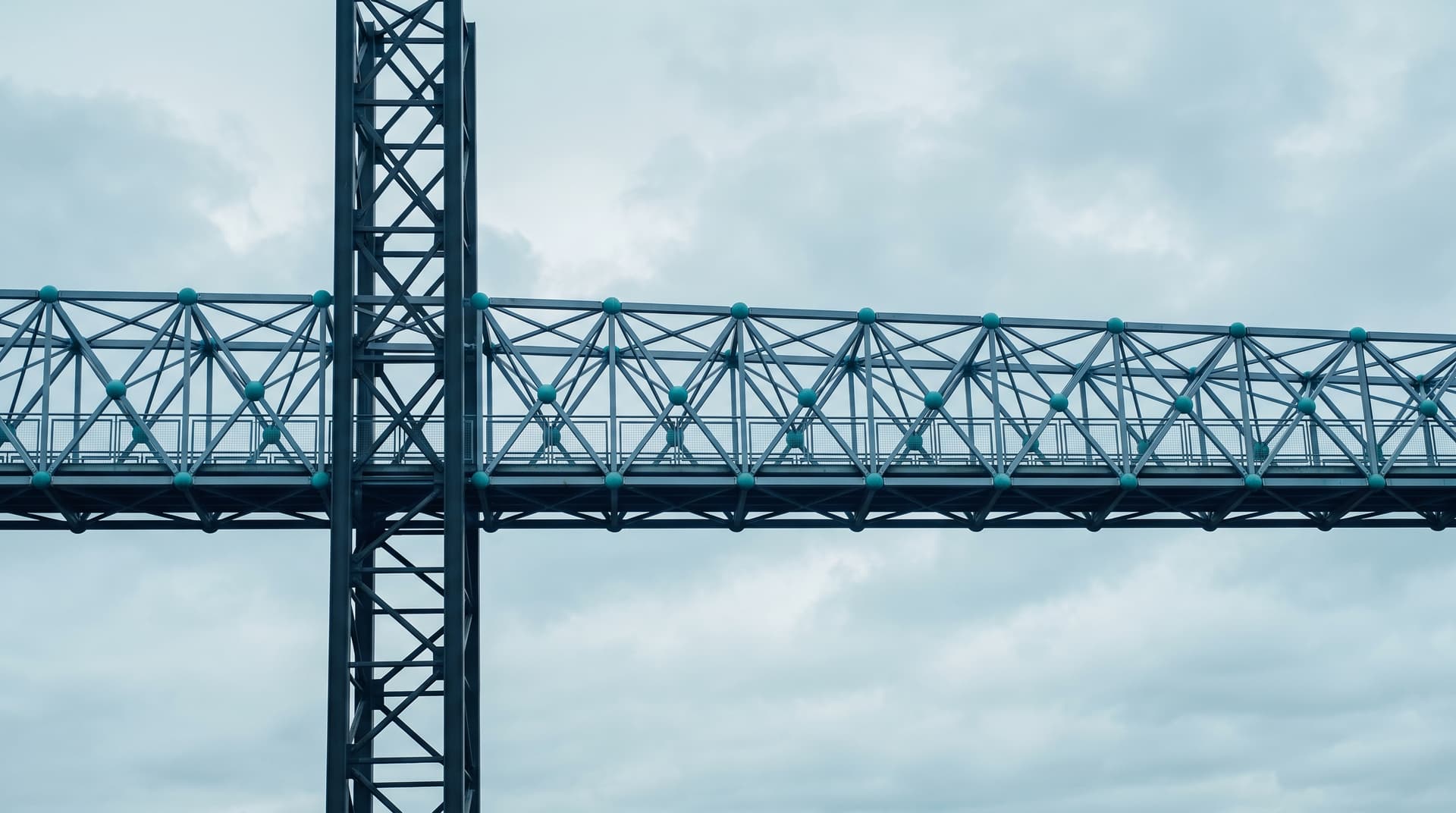 Stahlfachwerk-Fußgängerbrücke mit türkisfarbenen Knotenpunkten und senkrechtem Pylon vor bedecktem Himmel