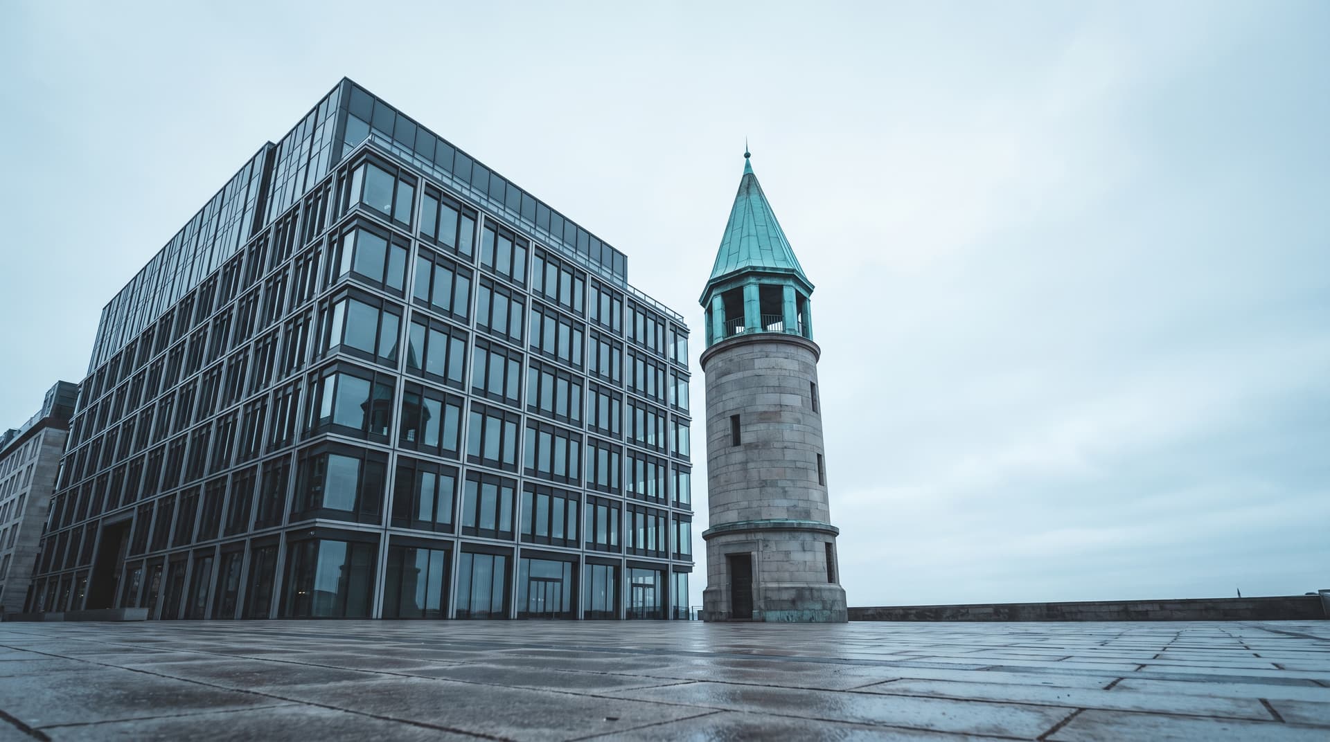 Großer Büroblock neben einem schmalen Turm mit patiniertem Kupferdach unter bedecktem Himmel, von unten fotografiert