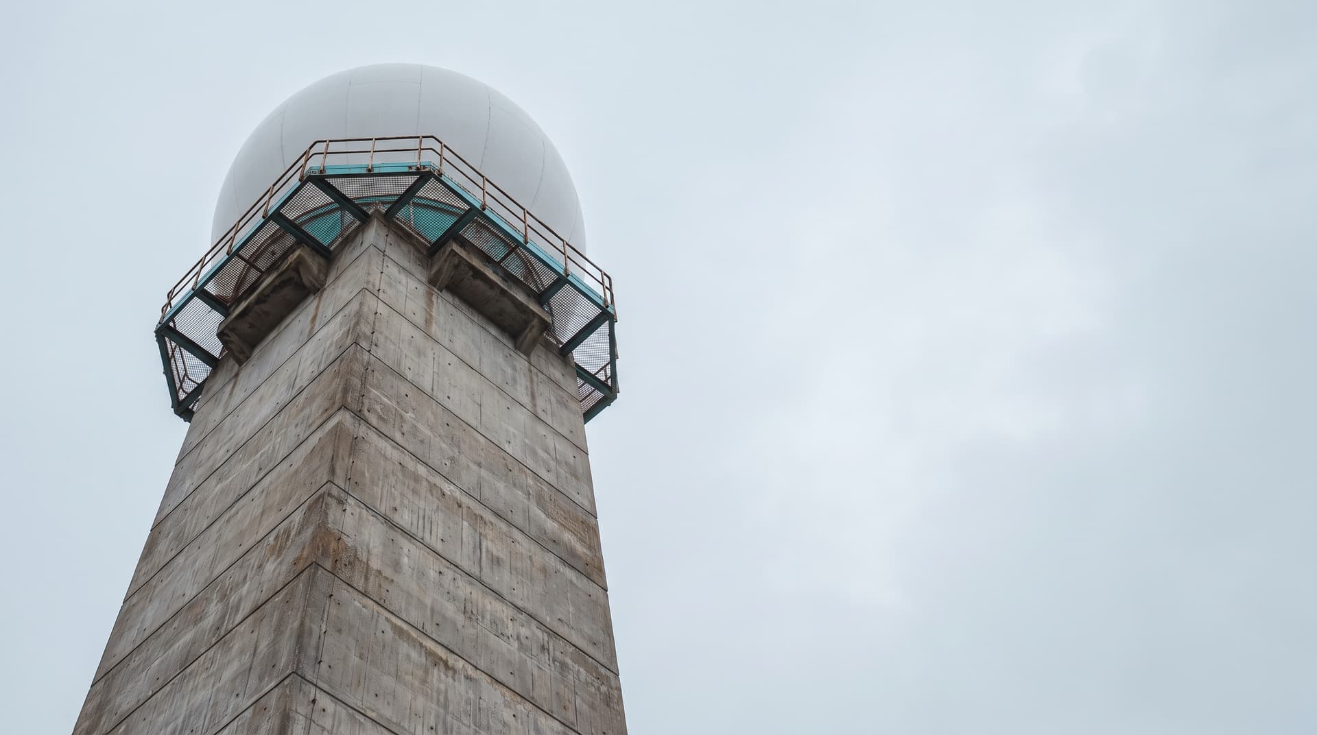 Concrete tower with a white radar dome photographed from below, platform railing in teal against a pale sky