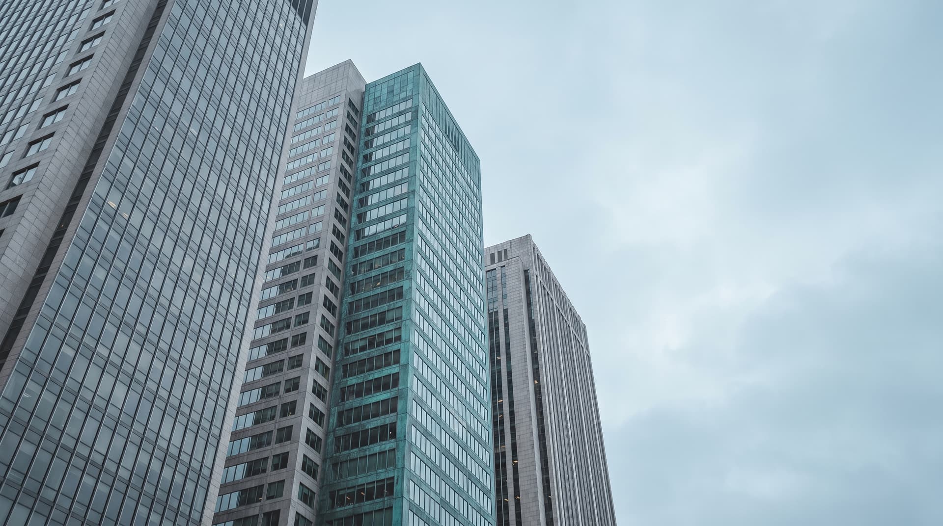 Gruppe von Bürohochhäusern, eines mit türkis getönter Glasfassade, unter bedecktem Himmel von unten fotografiert