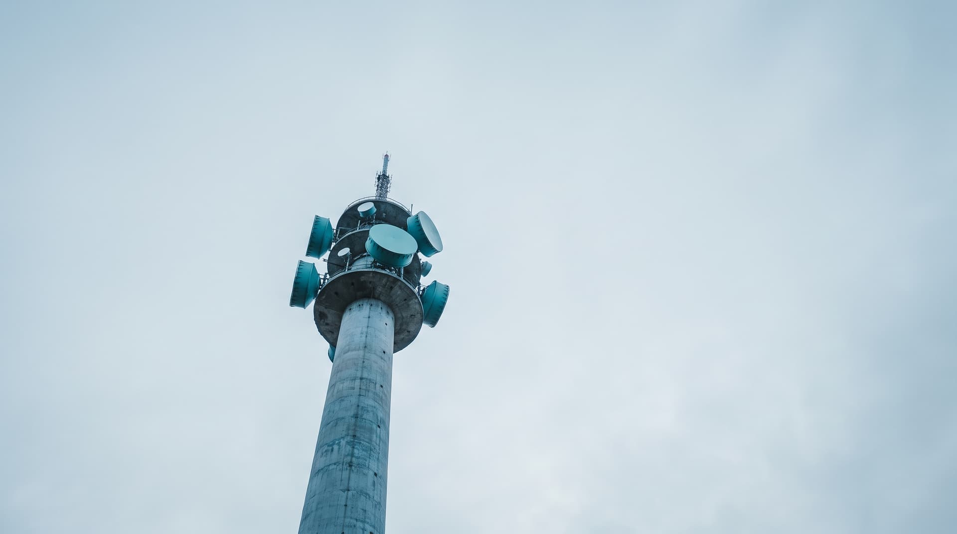 Tour de télécommunications élancée aux paraboles turquoise sous un ciel couvert, vue d'en bas
