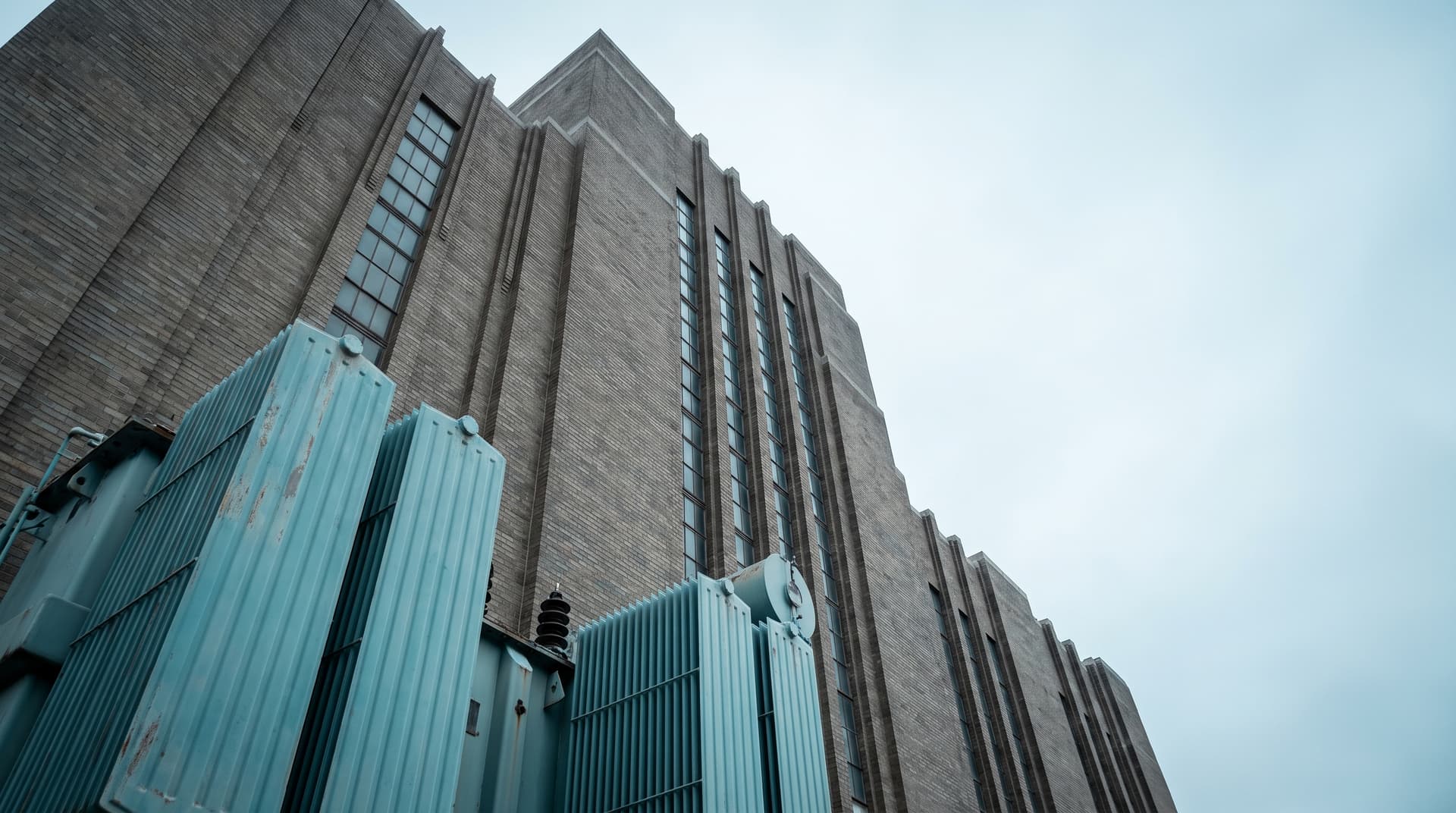 Historisches Backstein-Umspannwerk von unten mit hohen Fensterbändern, davor Transformatoren mit Kühlrippen in Teal – Sinnbild für Transformer-Modelle, die Energie in nutzbare Leistung umwandeln