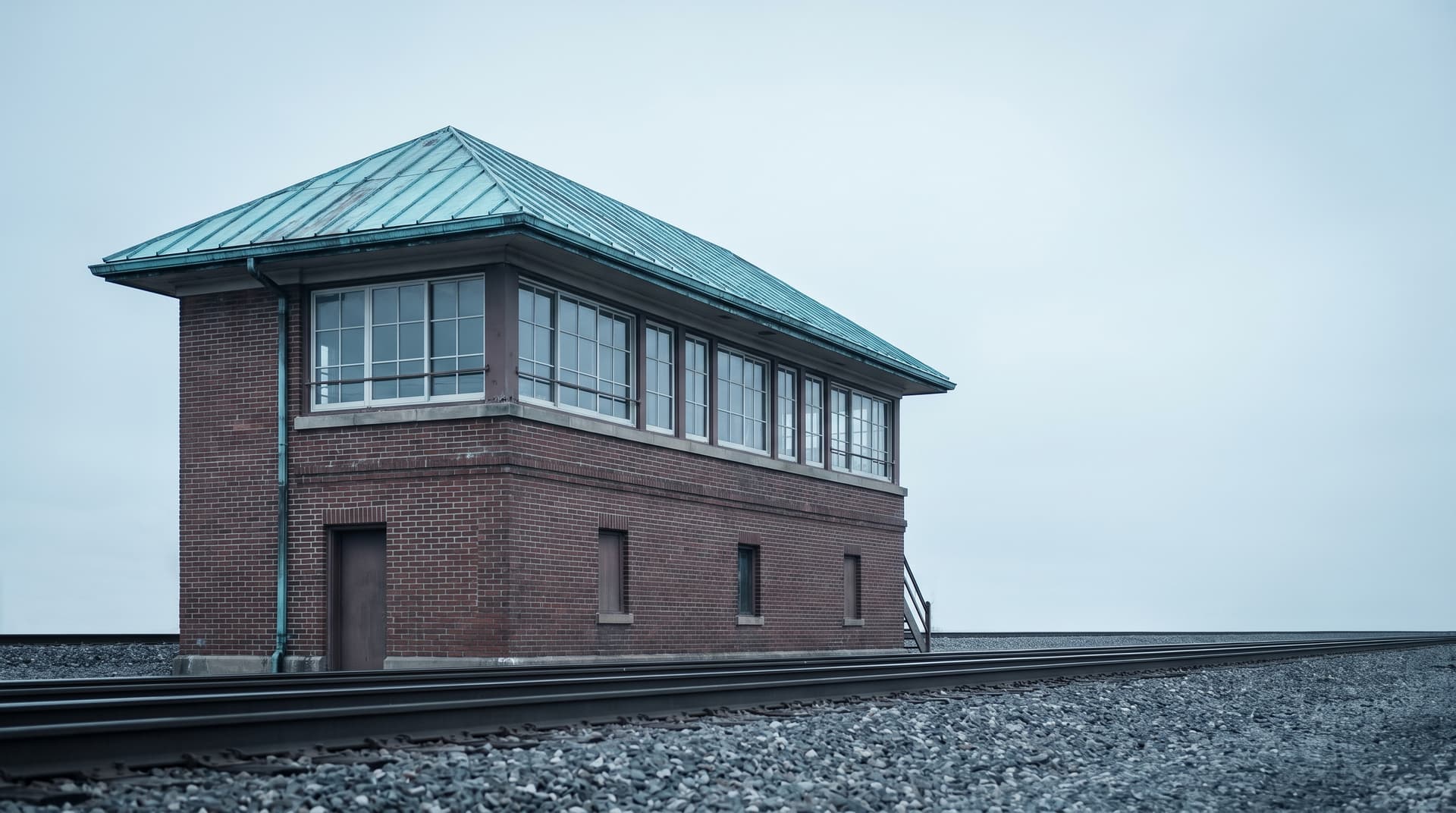 Brick railway signal box with a verdigris copper roof beside the tracks under an overcast sky – a metaphor for AI agents that control workflows