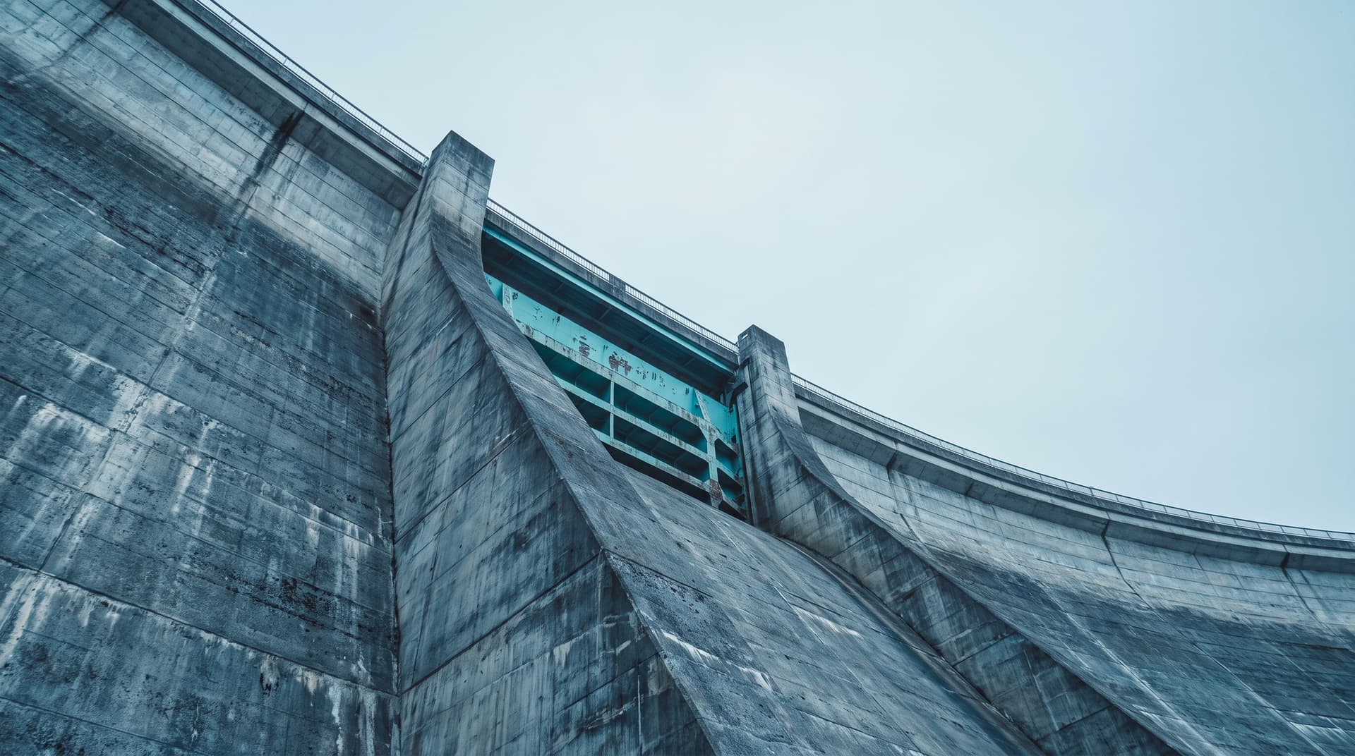 Betonstaumauer eines Wasserkraftwerks von unten fotografiert, Schützentor in Petrol als einziger Farbakzent