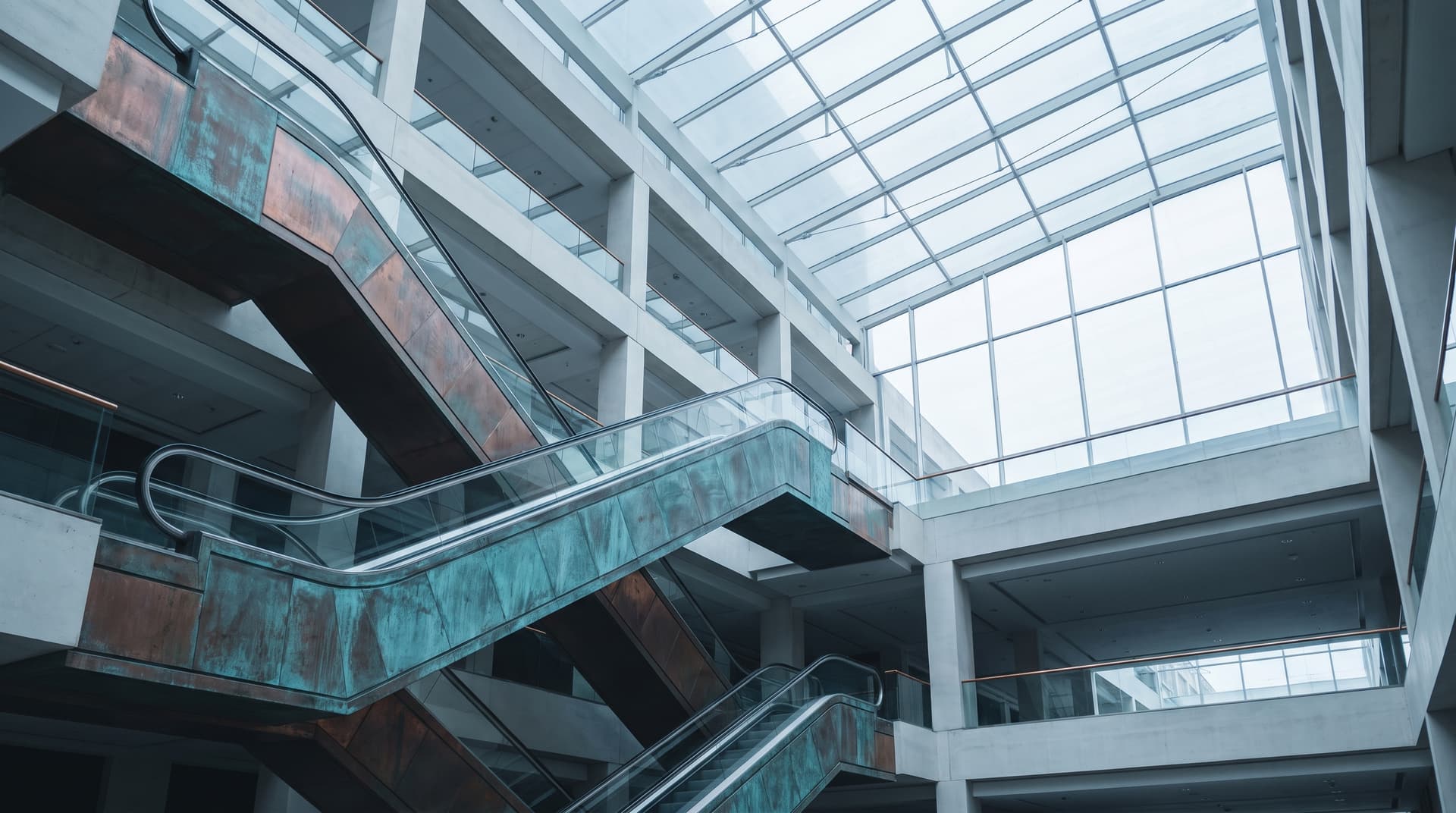 Crossing escalators with patinated copper panels in a shopping atrium under a glass roof, photographed from below