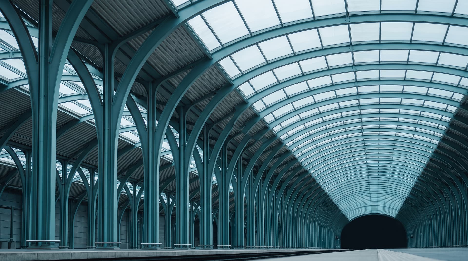 Stahl-Glas-Dach einer Bahnhofshalle mit türkis lackierten Stützen und Bögen, von unten fotografiert