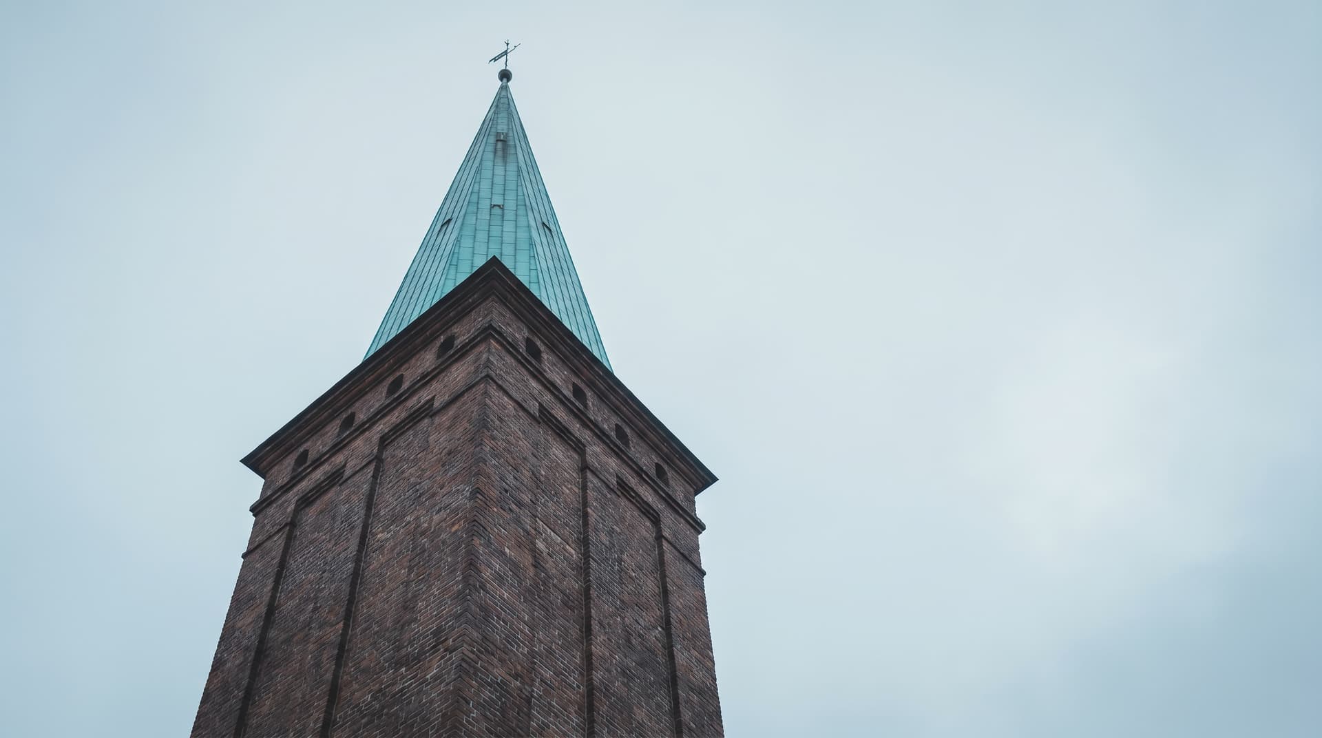 Brick tower with a pointed spire of green patinated copper and a weathervane, photographed from below