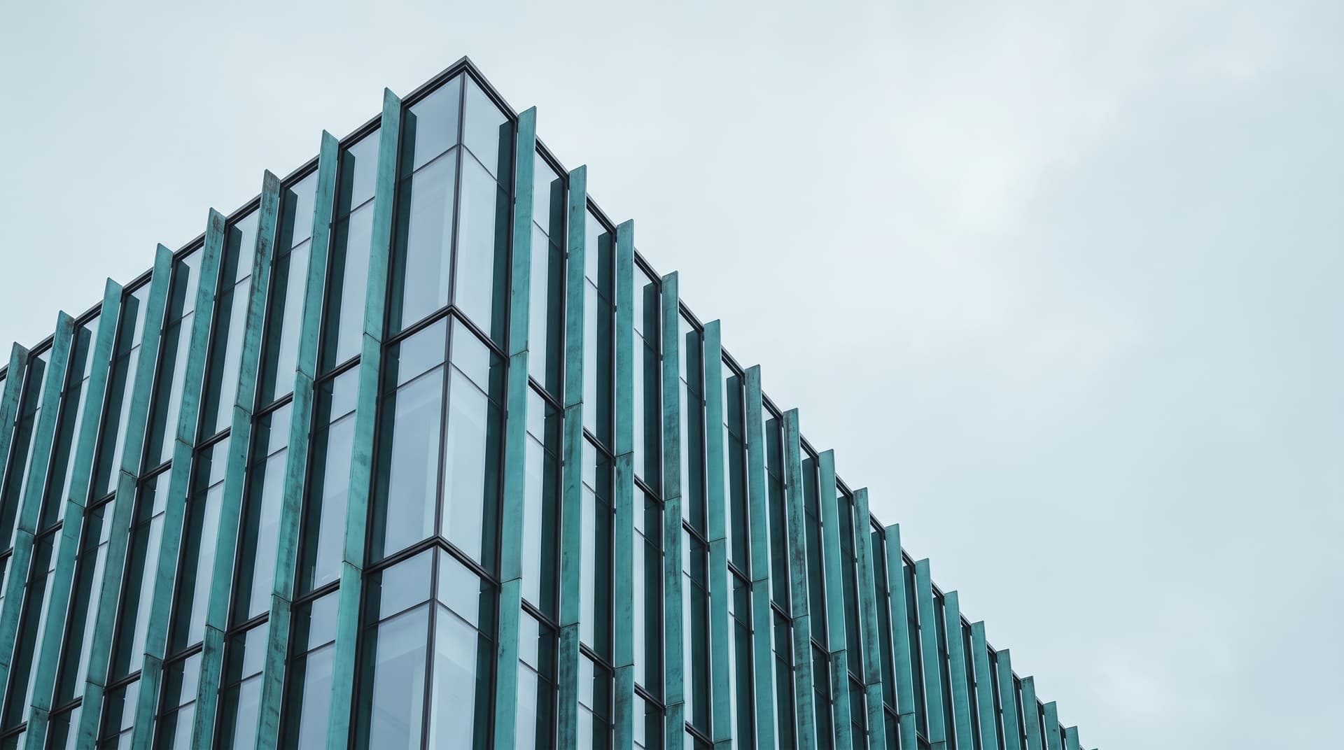 Gebäudeecke mit Glasfassade und senkrechten Lamellen aus patiniertem Kupfer unter bedecktem Himmel, von unten fotografiert